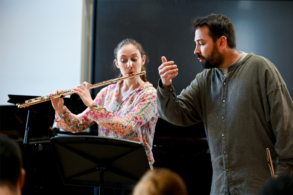 A man, with a dark beard, wearing a long-sleeved shirt, guiding a female student playing the flute.